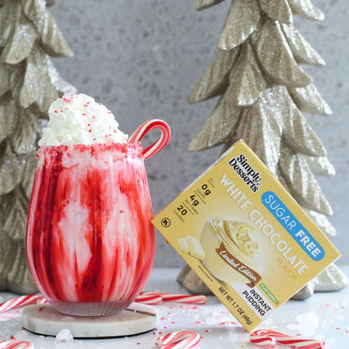 Festive Candy Cane Lane Mocktail topped with whipped cream, red sprinkles, and a candy cane, next to Simply Desserts White Chocolate Pudding box and gold holiday trees