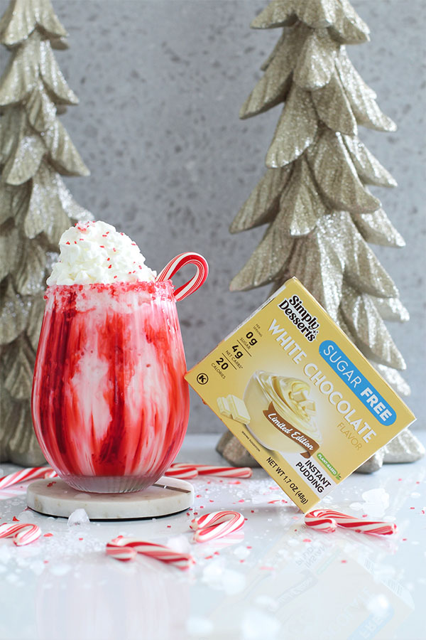 Festive Candy Cane Lane Mocktail topped with whipped cream, red sprinkles, and a candy cane, next to Simply Desserts White Chocolate Pudding box and gold holiday trees