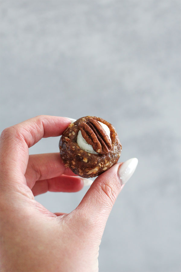 A close-up of a hand holding a White Chocolate Toffee Truffle, showing a rich date and pecan base filled with white chocolate pudding and topped with a pecan half