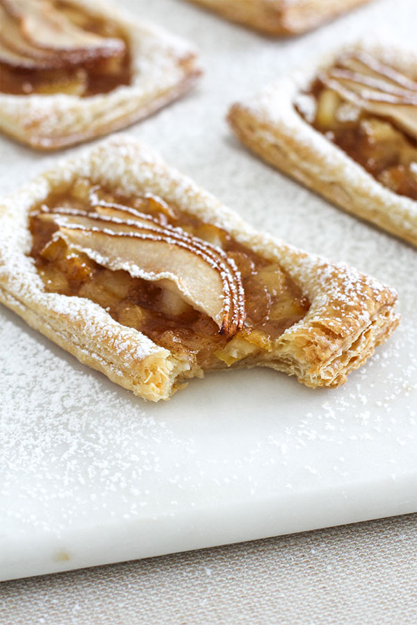 A close-up of a golden, flaky pear puff pastry tart with a bite taken out, dusted with powdered sugar on a white marble surface