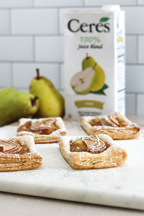 A set of pear puff pastry tarts displayed on a marble board, with a carton of Ceres Pear Juice Blend and fresh pears in the background