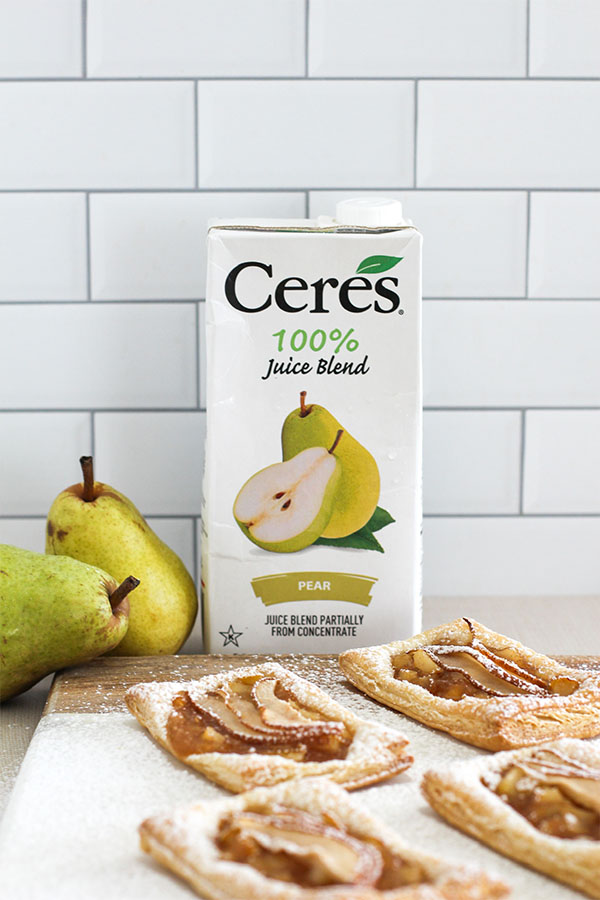 A carton of Ceres Pear Juice Blend with a batch of freshly baked pear puff pastry tarts on a white marble board, with whole pears nearby