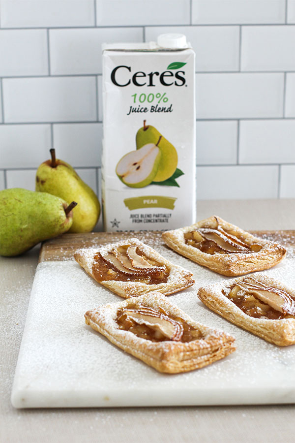 Four pear puff pastry tarts arranged on a marble board, with a blurred background featuring a Ceres Pear Juice Blend carton and whole pears
