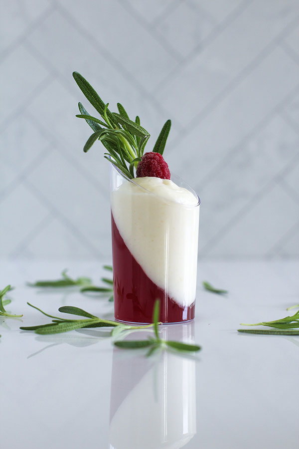Single white chocolate cranberry passionfruit shooter with a diagonal layer of cranberry-passionfruit jel, topped with a raspberry and rosemary sprig