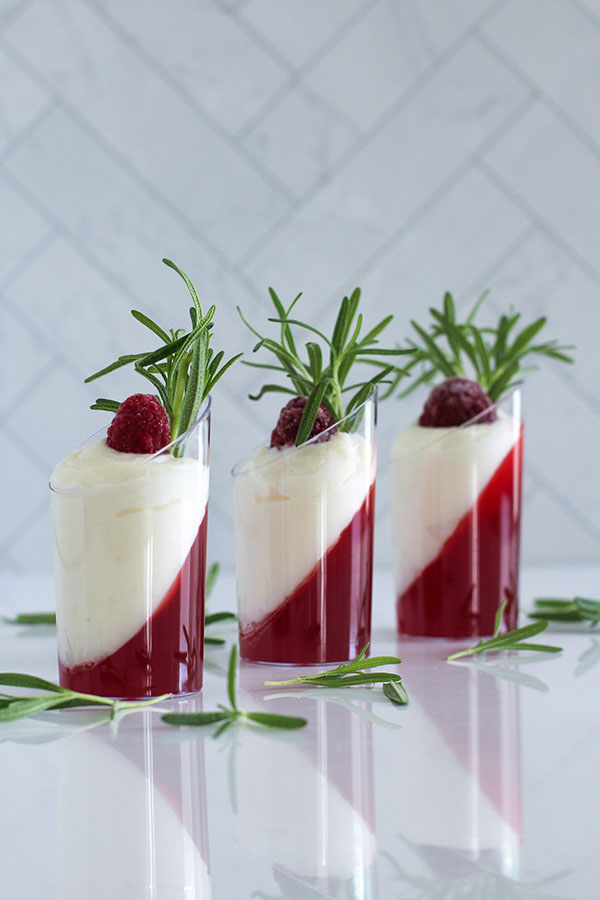 Three dessert shooters with a layered cranberry-passionfruit jel and white chocolate pudding, garnished with frozen raspberries and fresh rosemary