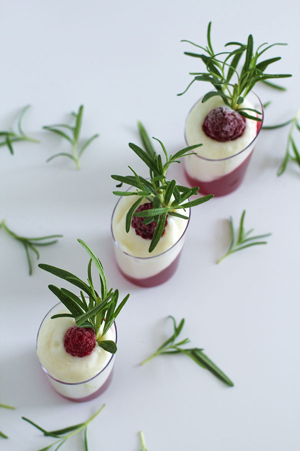 Overhead shot of three white chocolate cranberry passionfruit dessert shooters with rosemary and raspberry garnish, placed on a white surface with scattered rosemary sprigs