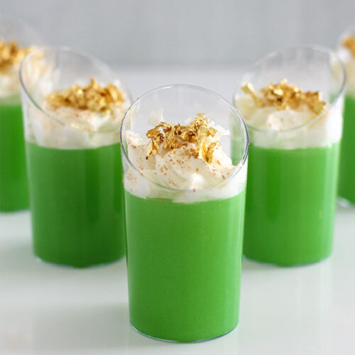 Close-up of green Irish Jello shots with whipped cream and gold flakes, arranged on a white surface