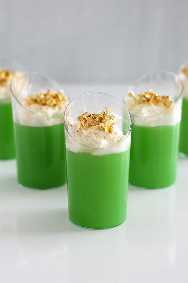 Close-up of green Irish Jello shots with whipped cream and gold flakes, arranged on a white surface