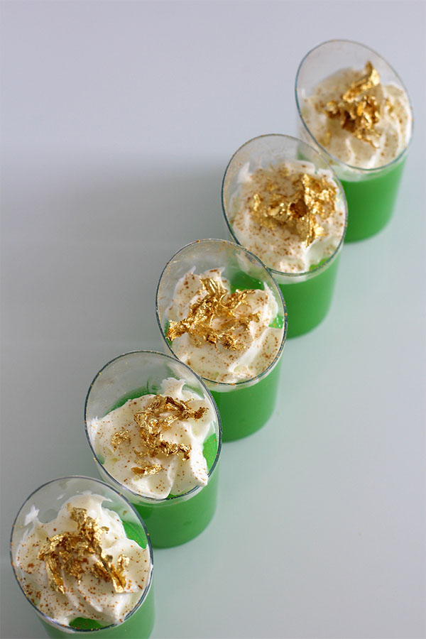 Diagonal row of green Irish Jello shots topped with whipped cream and gold flakes