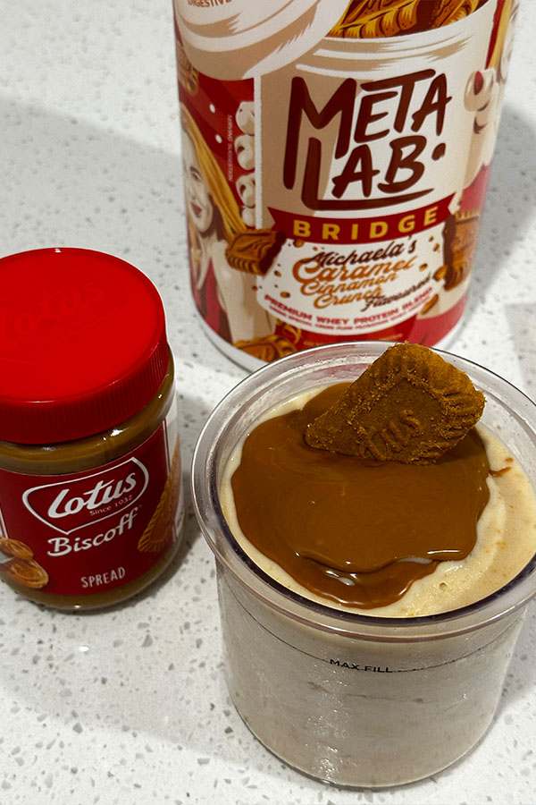 Biscoff protein ice cream in a Ninja Creami pint, topped with melted Biscoff spread and a biscuit. Lotus Biscoff spread jar and MetaLab protein powder tub are in the background