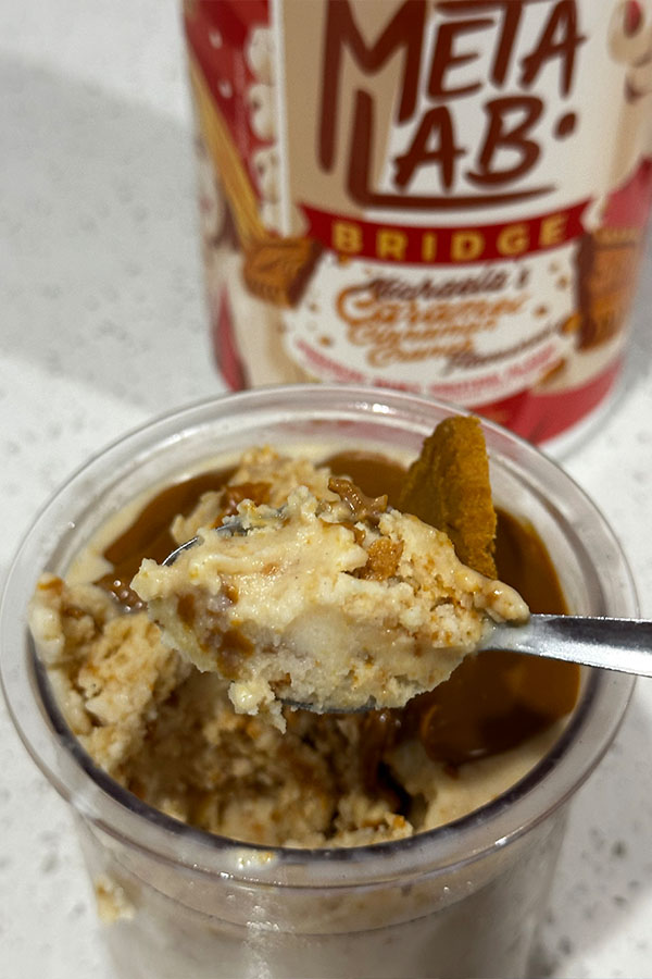 Close-up of a spoonful of creamy Biscoff protein ice cream showing biscuit chunks and spread, with the MetaLab protein tub slightly blurred in the background