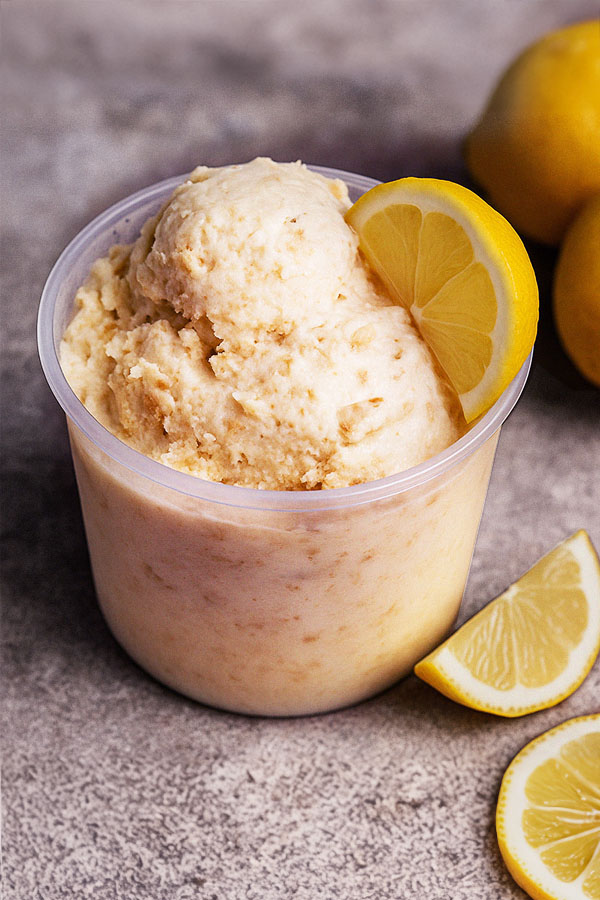 A clear plastic pint filled with lemon cheesecake protein ice cream, topped with a lemon slice. Slices of lemon and whole lemons are placed around it on a textured stone surface