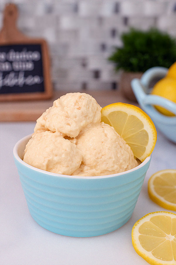 A single pastel blue bowl filled with smooth lemon cheesecake protein ice cream, topped with a lemon wedge. The background includes a chalkboard kitchen sign, plant, and bowl of lemons