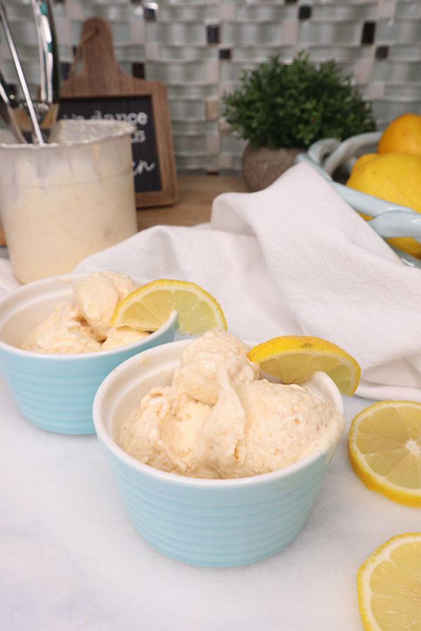 Two pastel blue ramekins filled with lemon cheesecake protein ice cream, each topped with a lemon slice. The background features a kitchen setting with a tub of ice cream, a white towel, a plant, and fresh lemons