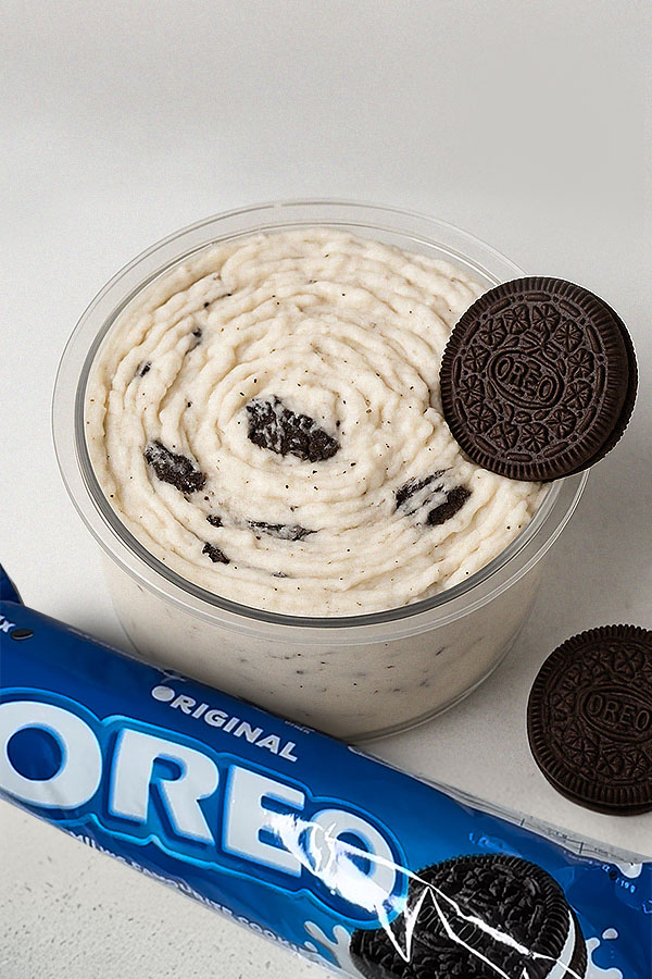 Oreo protein ice cream in a clear tub with cookie chunks, topped with an Oreo and a blue Oreo pack beside it.