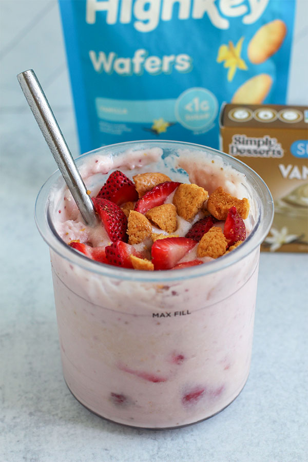 Ninja Creami pint filled with strawberry shortcake ice cream, topped with chopped strawberries and vanilla wafer pieces, placed in front of branded product packs.