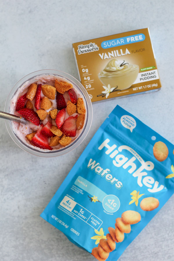 Flat lay of strawberry shortcake ice cream in a Ninja Creami container with Highkey Wafers and Simply Delish Vanilla Pudding packaging placed beside it.