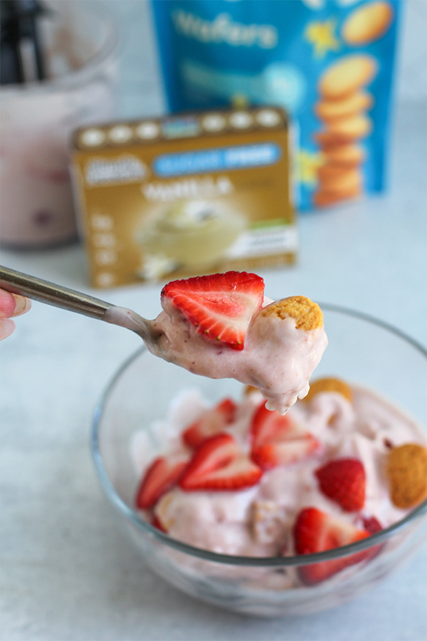Close-up of a spoonful of strawberry shortcake ice cream lifted above the bowl, with a strawberry slice and wafer on top, and product packaging softly blurred in the background