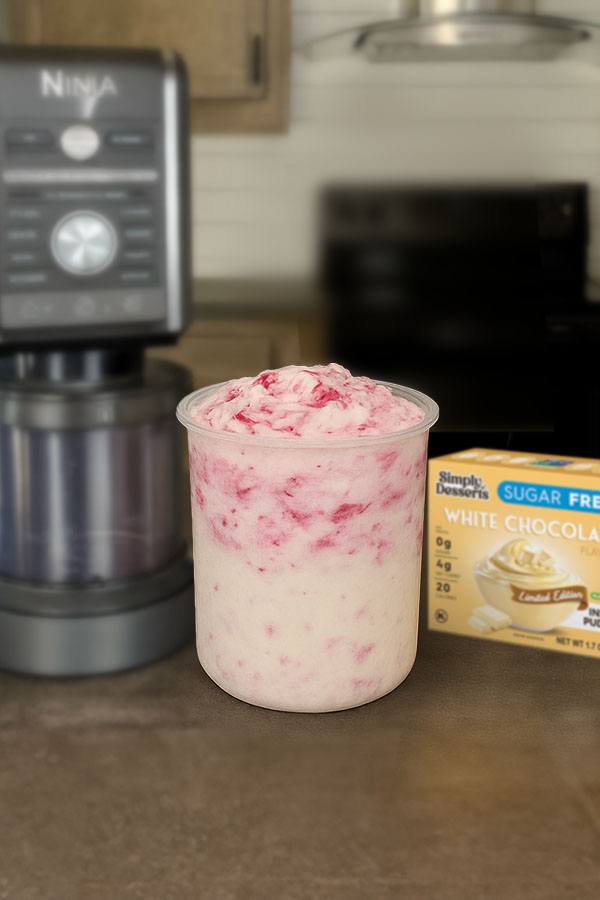 Freshly blended white chocolate raspberry cheesecake ice cream in a Ninja Creami pint container beside a Simply Desserts White Chocolate Pudding Mix box