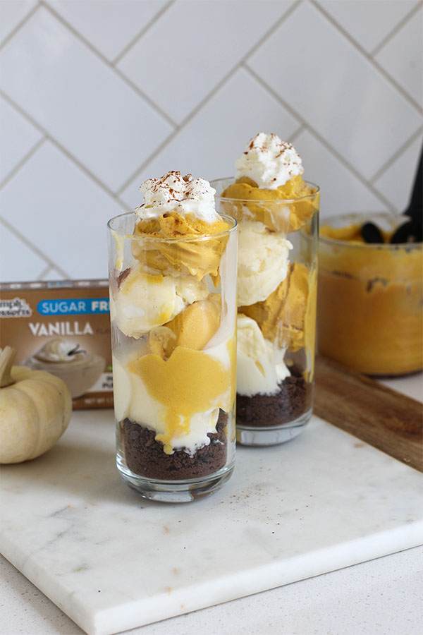 Two tall glasses layered with pumpkin and vanilla pudding over a chocolate crumb base, topped with whipped cream and cinnamon, displayed beside a Simply Desserts Vanilla Pudding box and a mini white pumpkin on a marble board