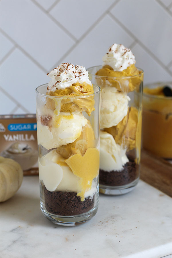 Two tall glasses filled with creamy layers of pumpkin and vanilla pudding over a chocolate crumb base, topped with whipped cream and sprinkled with cinnamon, with a Simply Desserts Vanilla Pudding box and fall decor in the background