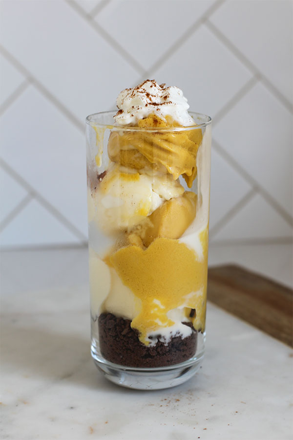 A tall glass layered with creamy pumpkin and vanilla pudding over a chocolate crumb base, topped with whipped cream and a sprinkle of cinnamon, creating a cozy fall-inspired dessert