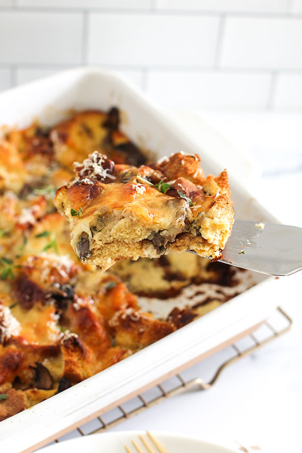 Golden savory bread pudding with mushrooms and melted cheese being lifted from a baking dish