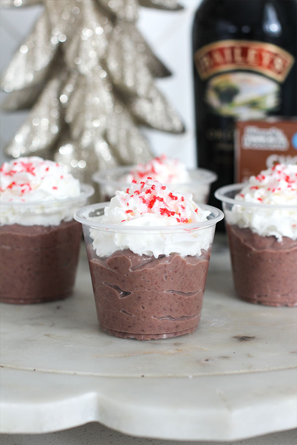 Creamy chocolate pudding shots topped with whipped cream and peppermint sprinkles with Baileys and Simply Desserts chocolate pudding in the background