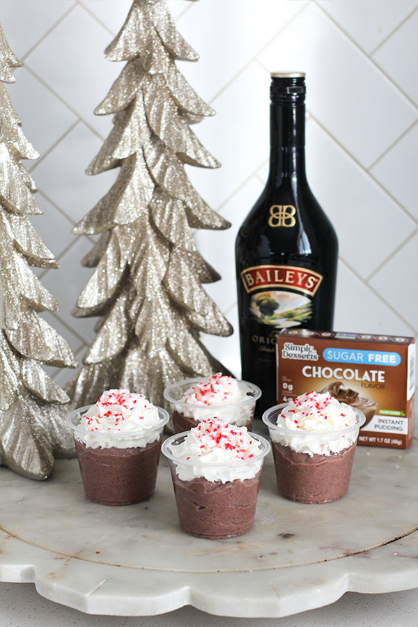 Four sugar-free hot chocolate pudding shots topped with whipped cream and peppermint sprinkles with a Baileys bottle and Simply Desserts chocolate pudding box behind