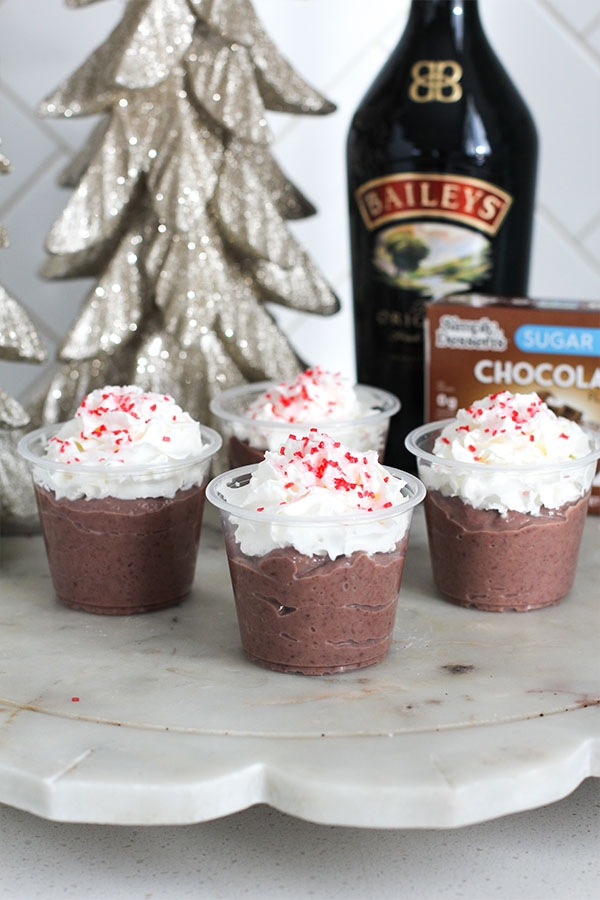 Row of chocolate pudding shots topped with whipped cream and peppermint sprinkles, styled with Baileys Irish Cream and Simply Desserts sugar-free chocolate pudding