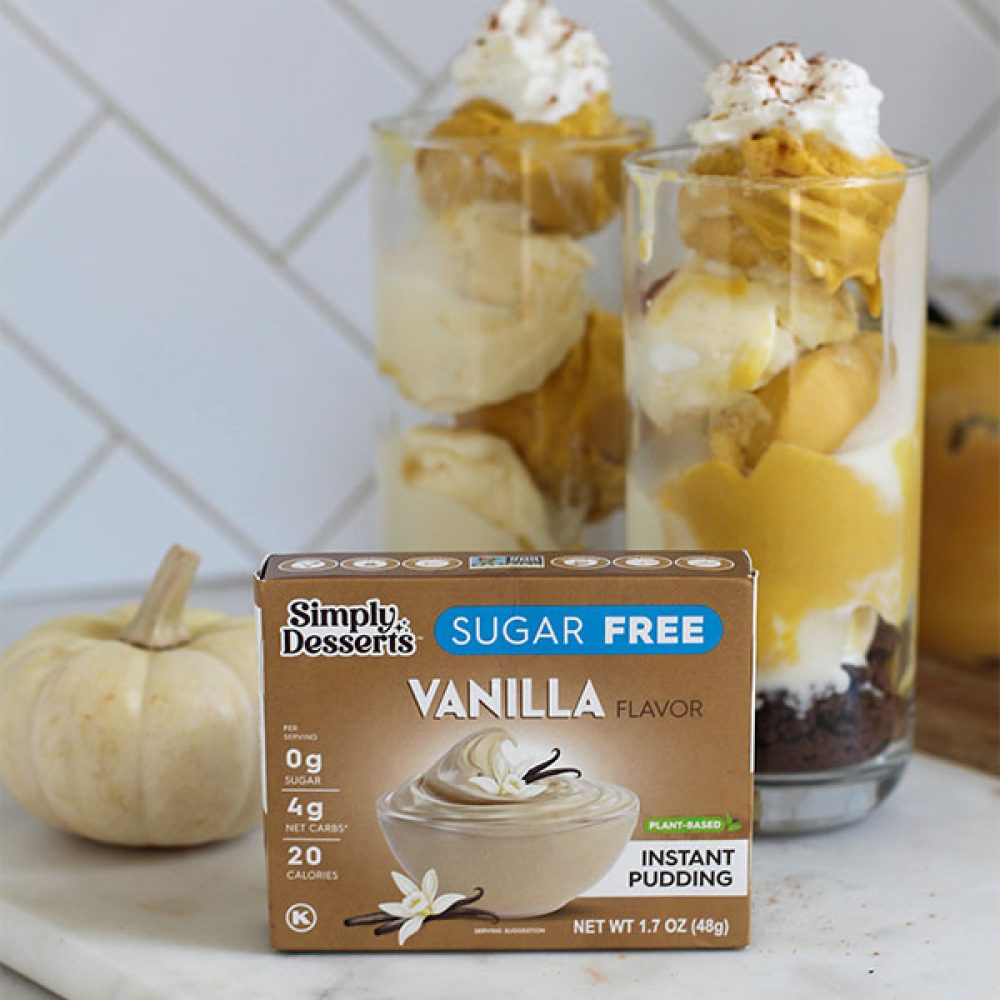 Simply Desserts Sugar-Free Vanilla Pudding box displayed on a marble board with two tall pumpkin and vanilla pudding sundaes topped with whipped cream and cinnamon in the background, styled with a small white pumpkin for a cozy fall look
