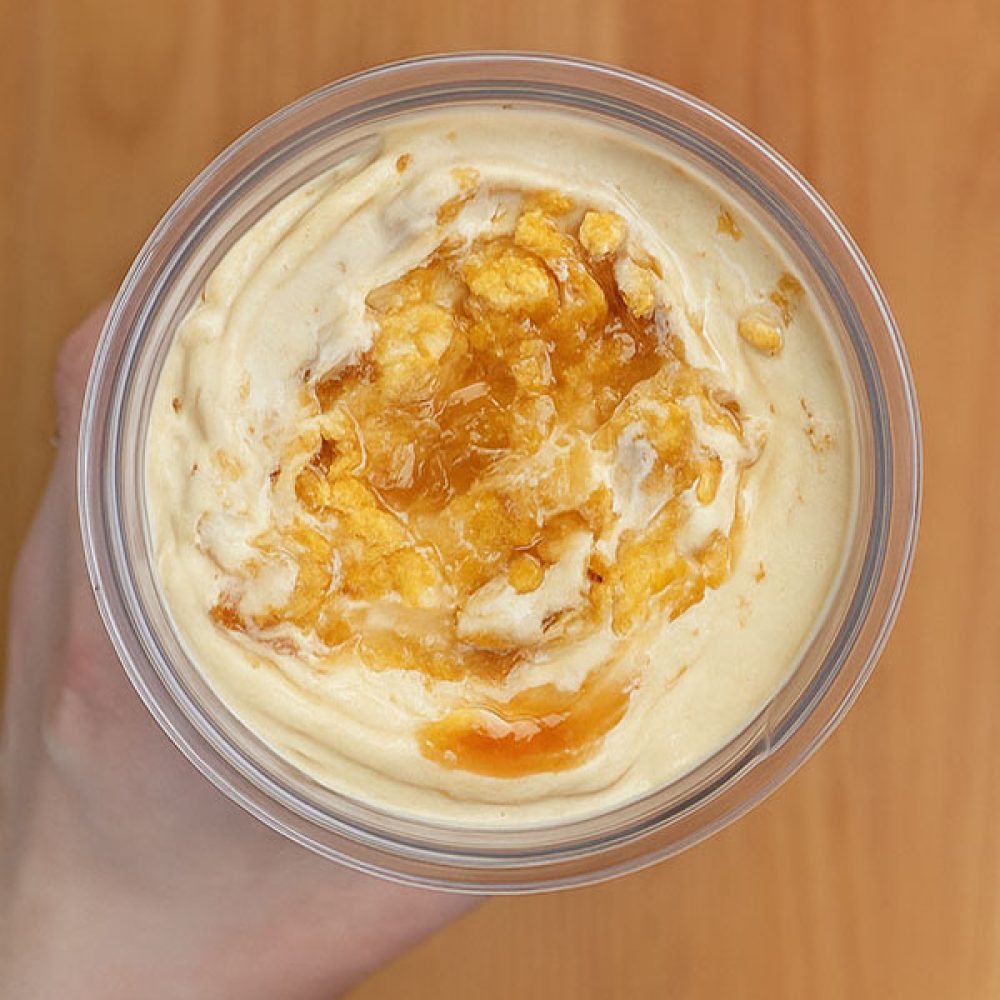 Top-down view of a hand holding a glass bowl of peanut butter caramel crunch protein ice cream, topped with golden caramel sauce and crushed cornflakes