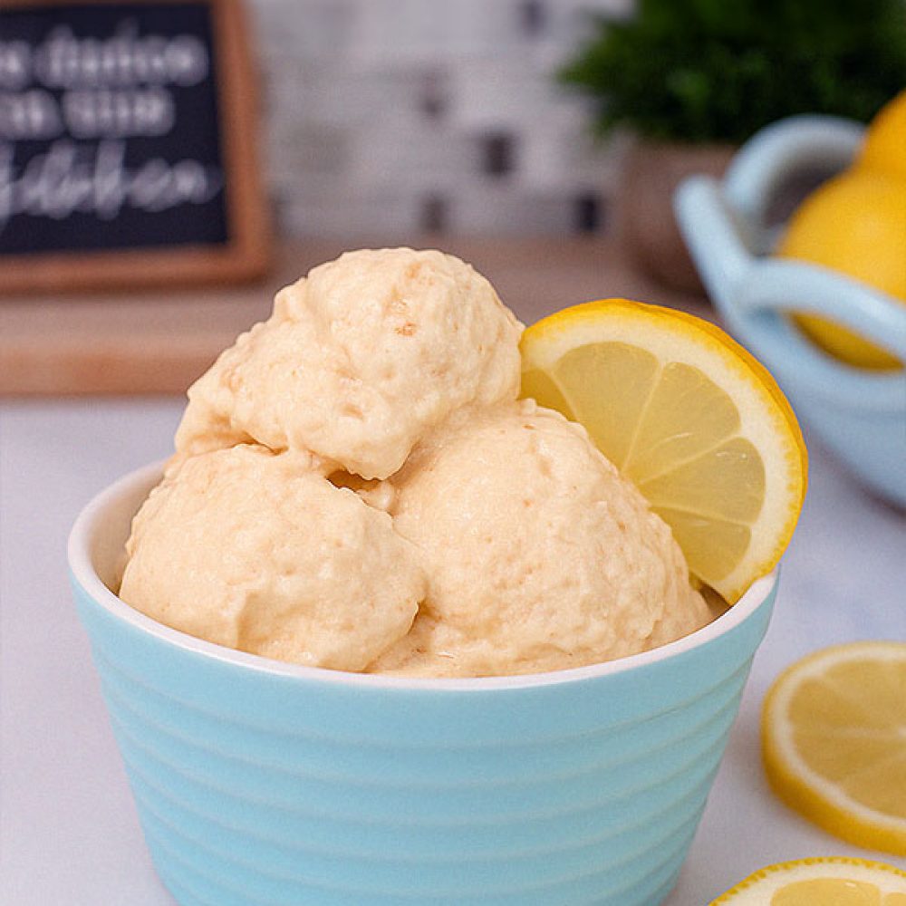 A single pastel blue bowl filled with smooth lemon cheesecake protein ice cream, topped with a lemon wedge. The background includes a chalkboard kitchen sign, plant, and bowl of lemons