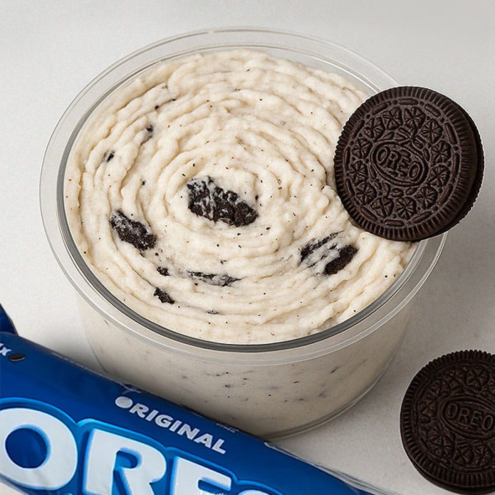 Oreo protein ice cream in a clear tub with cookie chunks, topped with an Oreo and a blue Oreo pack beside it.